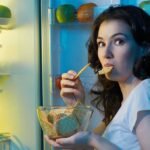 A woman eating a snack while looking at the camera standing in front of an open refrigerator