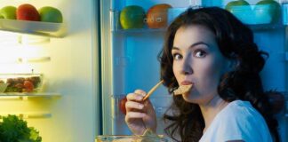 A woman eating a snack while looking at the camera standing in front of an open refrigerator