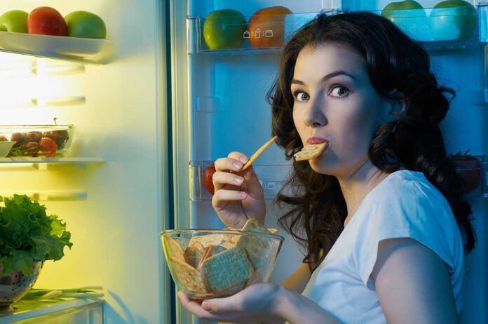 A woman eating a snack while looking at the camera standing in front of an open refrigerator
