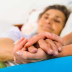 A caregiver holding the hands of an elderly patient in a hospital bed